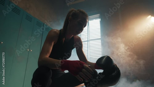 Serious woman fighter boxer wraps hands wrists with boxing bandage sits on bench in locker room, bottom view. Girl kickboxer prepares for fight. Professional sport, loss preparation concept. photo