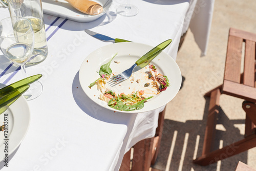 The remains of a summer salad on a white plate, with sun casting shadows over an outdoor table setting, signaling the end of a leisurely lunch. photo