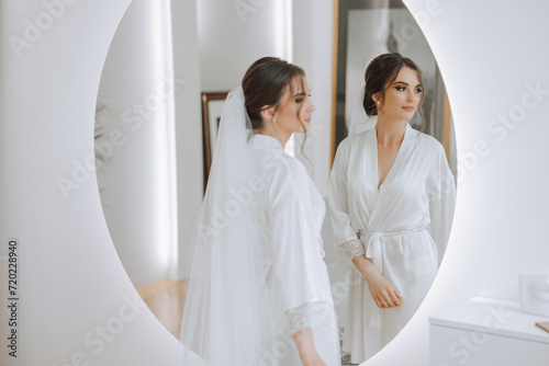 Bride in a wedding dress looking in the mirror. Beautiful young girl bride in the studio near the mirror in a white robe.