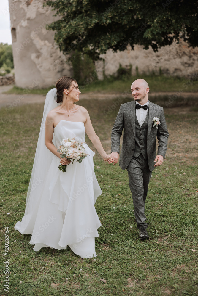 A handsome groom and an elegant bride in a lush white dress are walking in a summer park. Happy bride and groom getting ready for their best day.