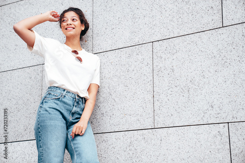 Young beautiful smiling hipster woman in trendy summer white t-shirt and jeans clothes. Carefree woman, posing in the street at sunny day. Positive model outdoors near wall. In sunglasses