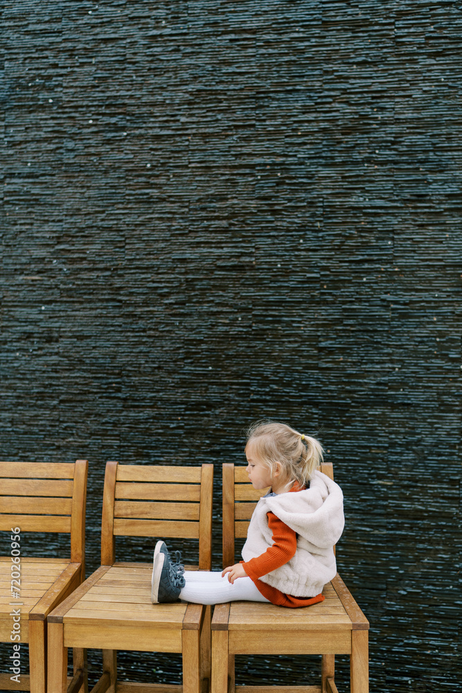 Little girl sits on a row of wooden chairs against the wall and looks at her shoes. Side view