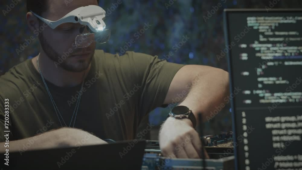 Waist up of Caucasian male military engineer in magnifying goggles with light assembling motherboard with programming code on computer display in focused foreground