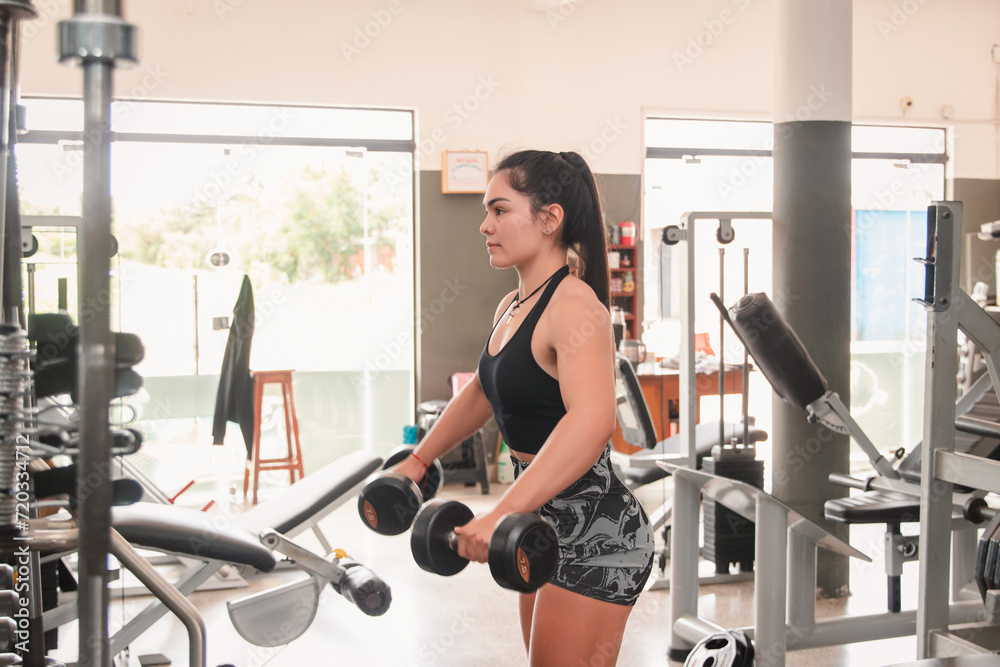 Fototapeta premium Fitness girl performing shoulder exercises with dumbbells in the gym.