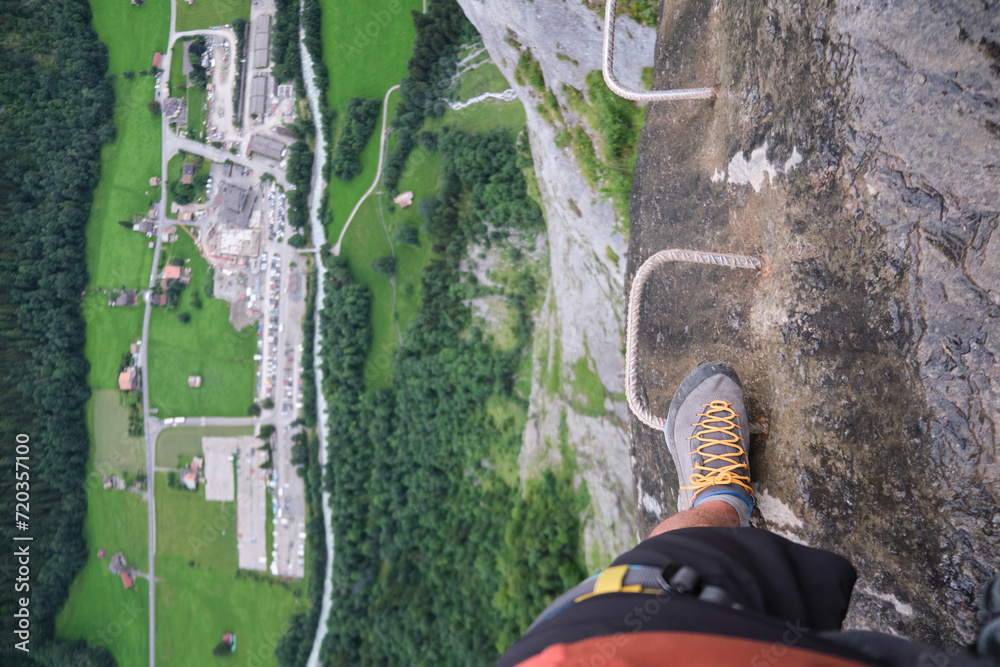 Tourist steps on metal via ferrata brackets, at the edge of a rock wall, with huge vertical gap under him and a view of the valley below. Summer activity at Murren-Gimmelwald klettersteig, Switzerland