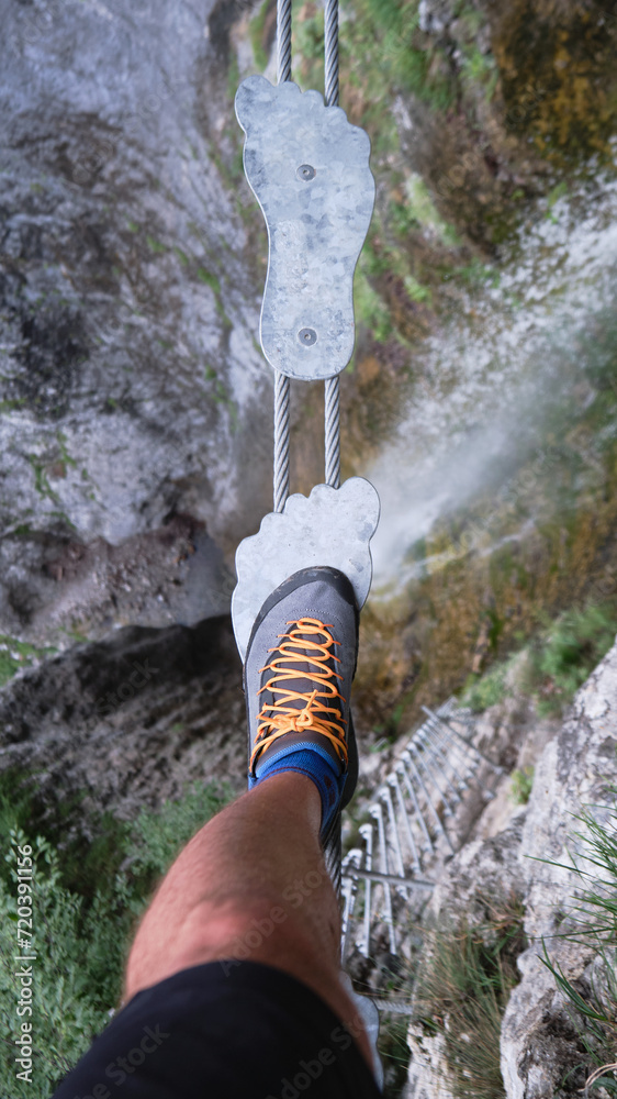 Tourist on foot shaped metal steps, on suspended bridge,over a waterfall, on via ferrata route in Italy, Europe. Summer adventure activities in the Dolomites mountains.