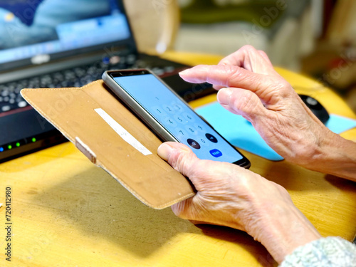 ノートパソコンの前でスマホの番号を操作する高齢女性の手元