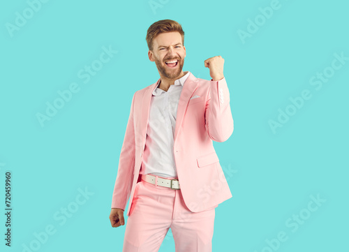 Portrait of a young overjoyed happy man with beard wearing stylish pink suit making yes winner's gesture celebrating success isolated on a studio blue background. People emotions concept.