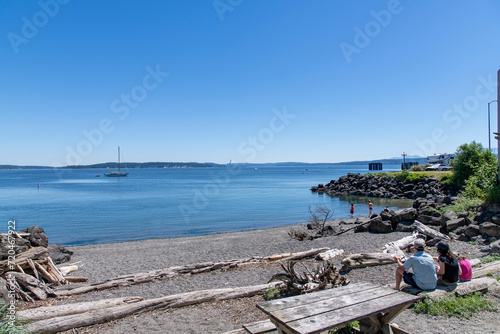 View over a small beach with people enjoying some lunch and others swimming on Tyler Street Plaza park in Port Townsend, WA, USA with views over the Port Townsend Bay against a clear blue sky