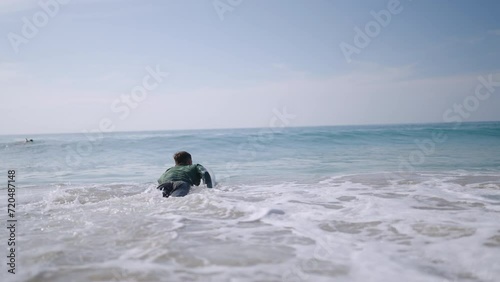 Male surfer in long sleeve rash guard paddling on surfboard. Man in rashguard swimwear swims to lineup lying on board. Surfer goes to ocean water swells. Recreational activity and water sport.