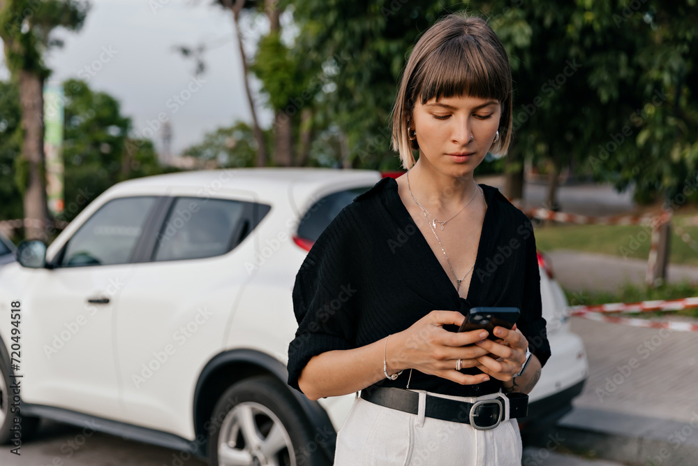 © PhotoBook - Stylish pretty girl with short hair wearing black shirt is using smartphone on background of the car © PhotoBook - Stylish pretty girl with short hair wearing black shirt is using smartphone on background of the car
