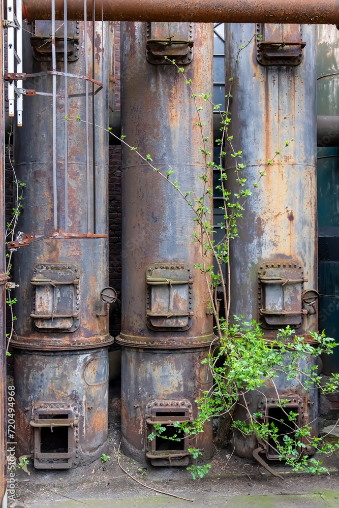 Three rusted and weathered furnaces or piping systems with steel doors of an abandoned and dilapidated industrial steel mill in public Landschaftspark, Duisburg with some green growing in front