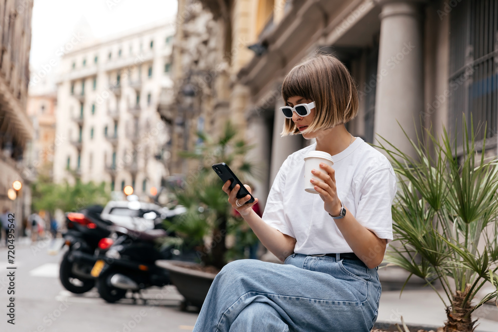 Stylish modern girl with short hair is looking at smartphone and enjoying coffee outdoor on background of historical city