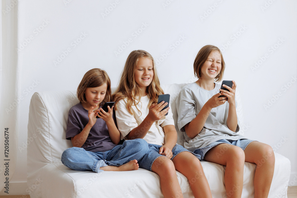 Three girls use smartphones on the sofa. Three sisters smiling in casual clothes against a white wall. Concept for advertising a mobile application or content for the whole family. Three girls use smartphones on the sofa. Three sisters smiling in casual clothes against a white wall. Concept for advertising a mobile application or content for the whole family.
