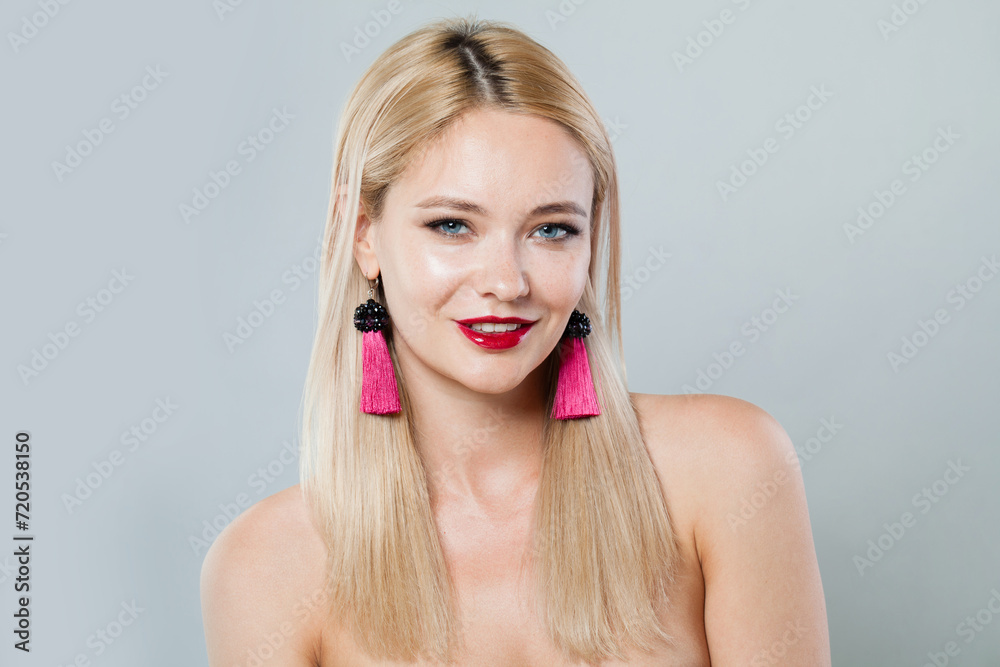 Obraz premium Happy blonde woman with fashion jewelry eraring, blond hairstyle, clean fresh skin and red lipstick make-up on gray background, studio portrait