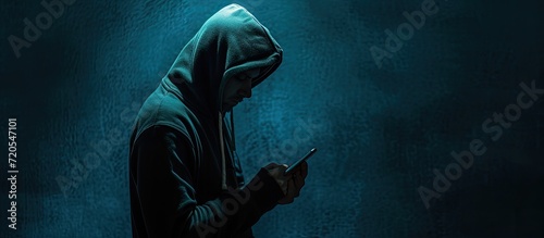 The image portrays a shadowy hacker, concealed by a hood, brandishing a smartphone, embodying the sinister world of cybercrime, internet breaches, and malware attacks against a dark background.