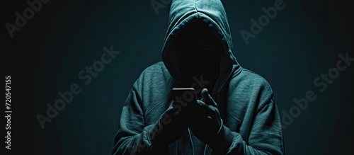 The image portrays a shadowy hacker, concealed by a hood, brandishing a smartphone, embodying the sinister world of cybercrime, internet breaches, and malware attacks against a dark background. photo