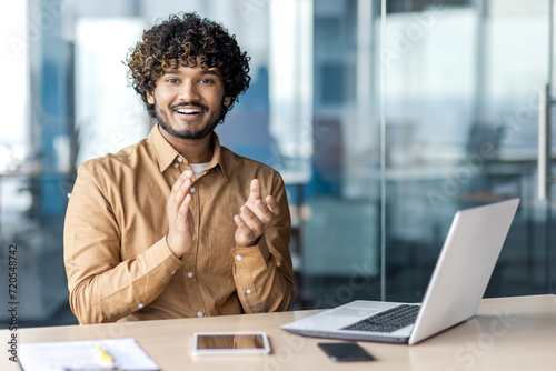 Joyful indian man in casual wear clapping hands while sitting at workplace with laptop in modern office. Young male with curly hair congratulating colleague with successful project and smiling.