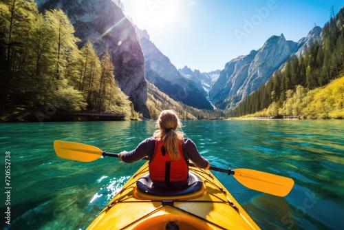 Person kayaking in a vibrant mountain lake surrounded by lush forests and majestic peaks on a sunny day. © Thanaphon