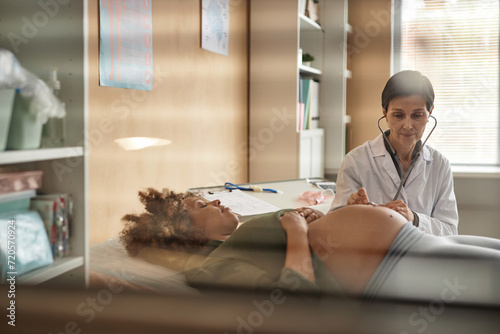 Look through ward window of friendly mature Caucasian gynecologist with stethoscope checking fetal heart tones while pregnant Black female patient lying down during clinic visit photo