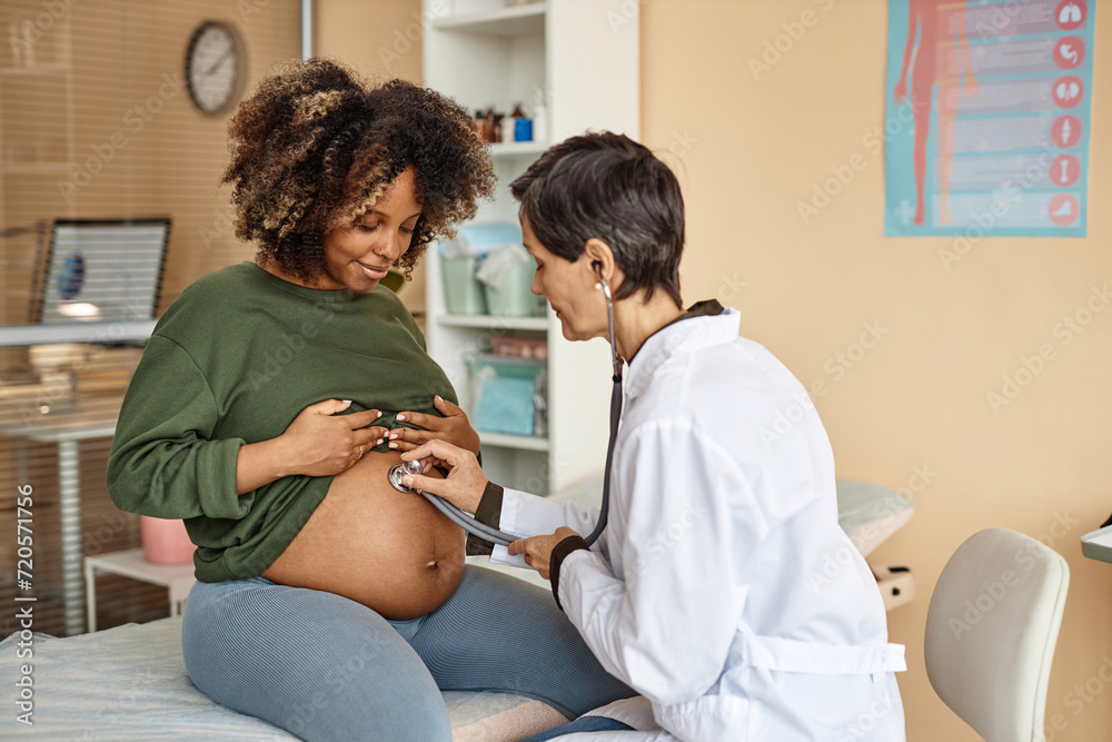 © Seventyfour - Smiling young African American expectant mother sitting on medical table in clinic while female obstetrician using stethoscope listening baby © Seventyfour - Smiling young African American expectant mother sitting on medical table in clinic while female obstetrician using stethoscope listening baby