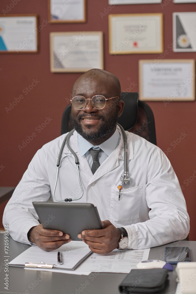Black medical worker sitting at his desk in clinic with tablet in his hands and looking at camera