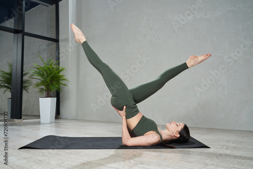 Young slender woman in tight leggings practicing yoga legs up pose on yoga mat at home. Side view of focused girl doing hatha yoga postures in modern indoor space. Concept of yoga practice, sport.  photo