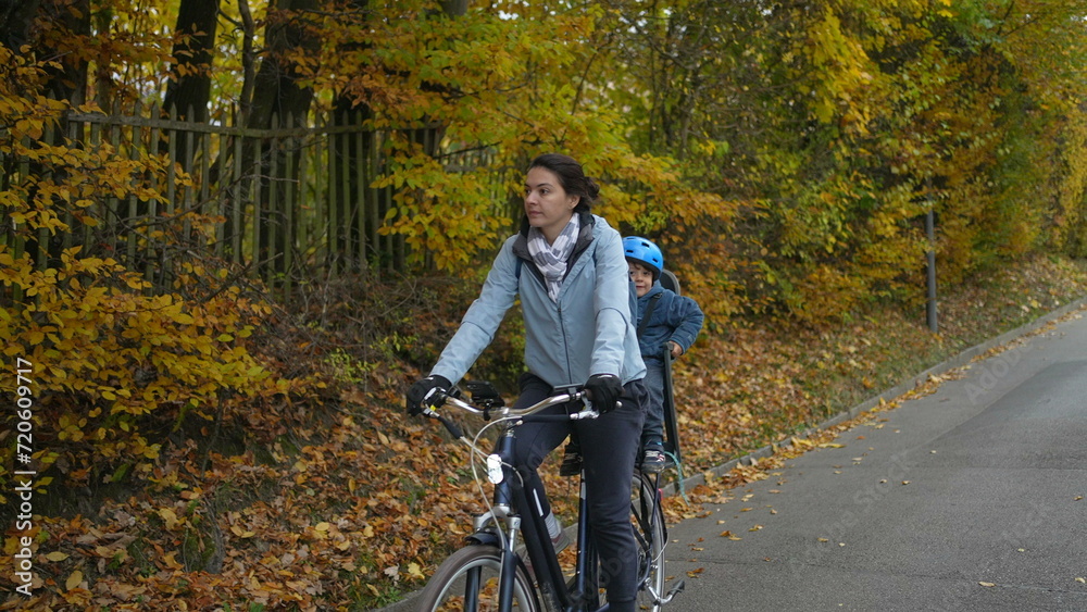Fototapeta premium Mother rides bicycle with child in back seat during autumn fall day. Parent riding bike with son wearing helmet and jacket