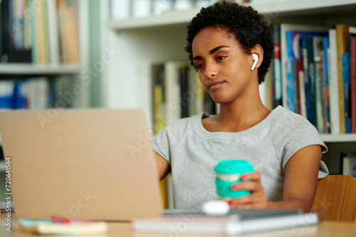 Black woman is immersed in her work on a laptop, holding a reusable coffee cup, showcasing a sustainable and attentive approach to her remote professional life.