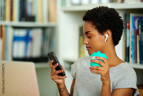 young black woman simultaneously manages tasks on her smartphone with earbuds in and enjoys a drink from an eco-friendly cup, embodying modern efficiency. photo