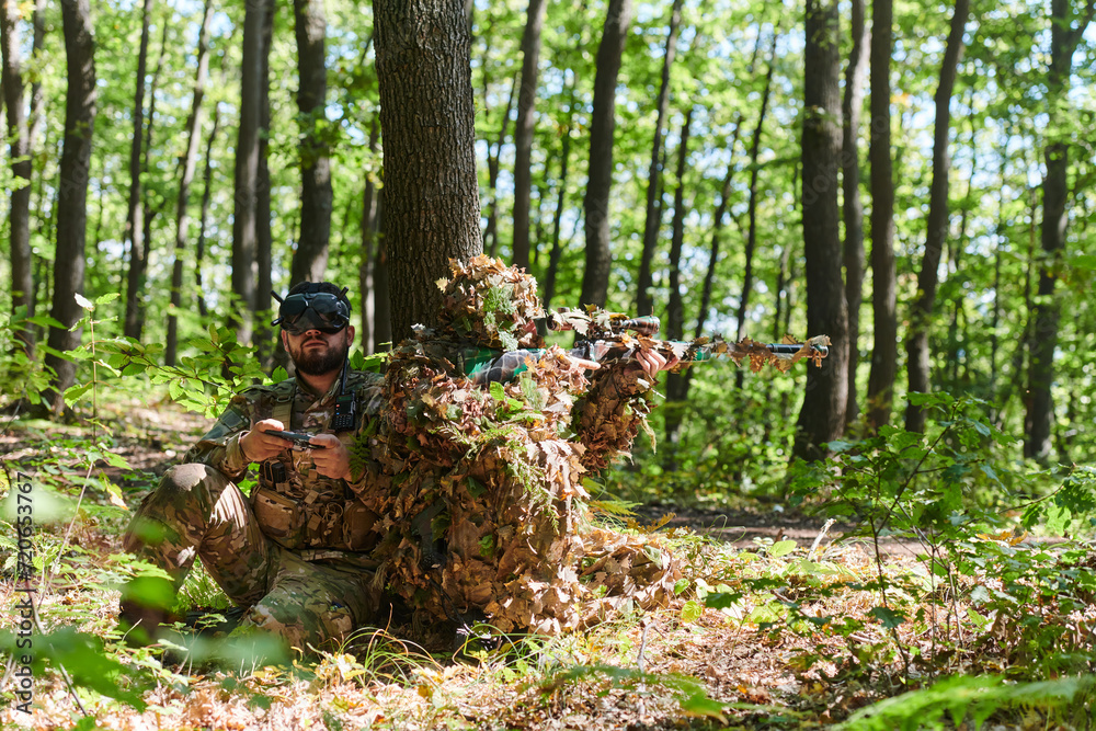 A skilled sniper and a soldier operating a drone with VR goggles strategize and observe the military action while concealed in the forest