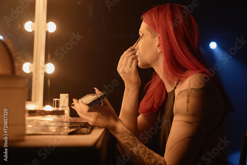 Profile view of of tattooed young woman putting on makeup and applying eyeshadow with brush in dressing room before dance show