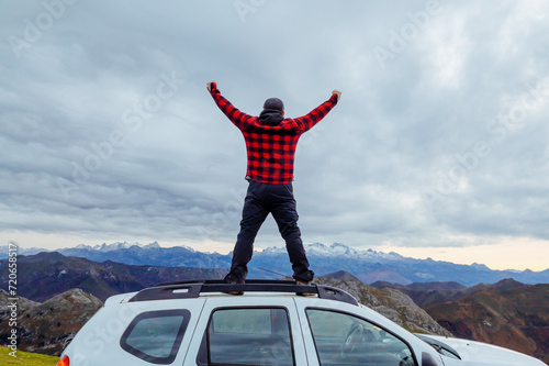 man standing on the roof of his 4x4 SUV car with his arms outstretched looking towards the mountains. offroad adventure trip