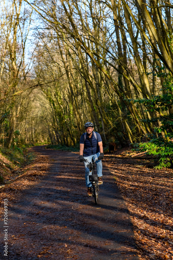 Senior cyclist rides along a shared bicycle path through woodland. He's wearing a bike helmet riding an electric folding bike. A retirement wellness exercise and lifestyle capture. copy space.