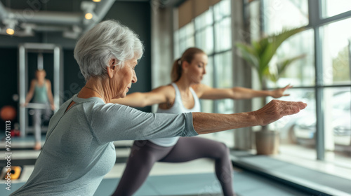 elderly woman practicing yoga with her arms extended, focusing intently, with another person in the background in a bright, window-lined room