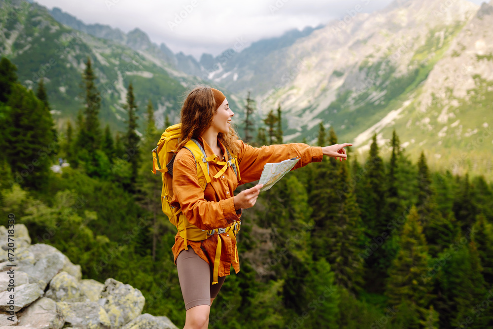 Young woman traveler with a yellow backpack, holds a map, explores hiking trails in the mountains. The concept of travel, vacation.  Hiking and adventure concept