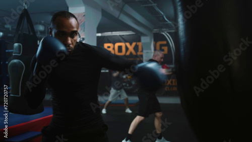 African American fighter listens coach command and starts hitting punching bag in dark boxing gym. Athletic man in boxing gloves exercises before tournament. Physical activity and intensive workout.