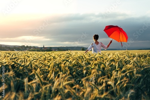 Junge Frau mit einem roten Schirm steht in einem Getreidefeld und schaut Richtung Sonnenuntergang. Ein Kirchturm am Horizont.