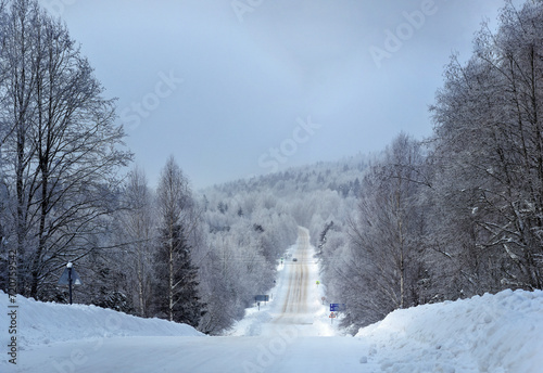 Winter mountain rural landscape in the afternoon in cloudy weather. A road through a snow-covered meadow among a spruce forest on a hill