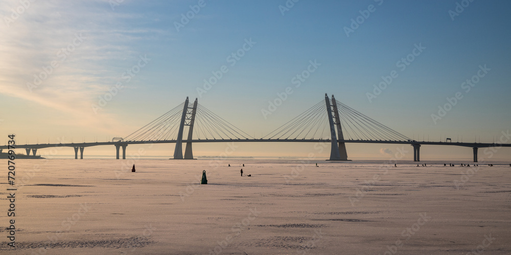 View of the cable-stayed bridge over the Ship Fairway in St. Petersburg in winter.The Neva Bay of the Gulf of Finland is covered with ice, there are many fishermen on the ice engaged in winter fishing View of the cable-stayed bridge over the Ship Fairway in St. Petersburg in winter.The Neva Bay of the Gulf of Finland is covered with ice, there are many fishermen on the ice engaged in winter fishing