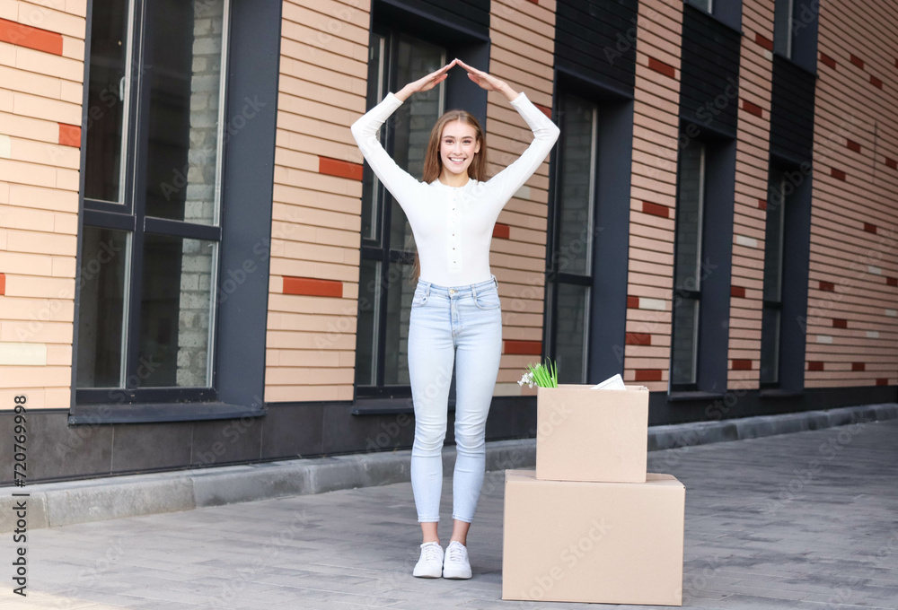 Pretty young woman is standing near box with decor for home and at inner yard of apartment building. She is relocating to a new home. She is happy. She is showing the house with her hands. Pretty young woman is standing near box with decor for home and at inner yard of apartment building. She is relocating to a new home. She is happy. She is showing the house with her hands.