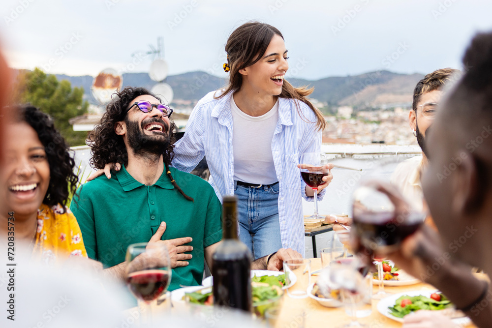 Group of diverse young adult friends enjoying having lunch together in a terrace with city views. Happy friends social gathering and having fun during barbecue at rooftop party.