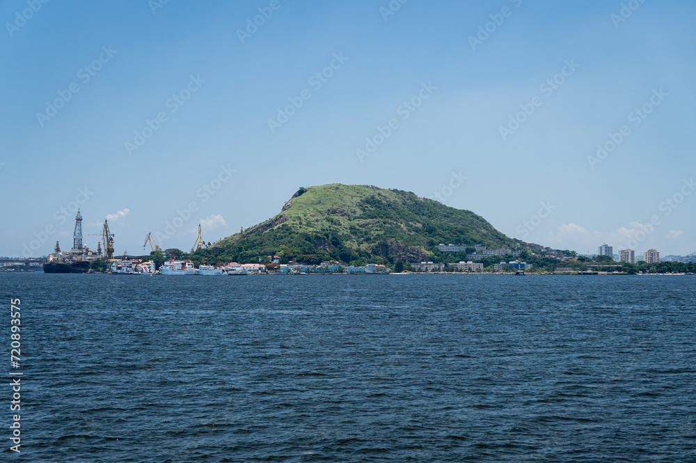 Distant view of Morro da Penha hill in Niteroi's Ponta Dareia district as saw from Guanabara bay blue waters under summer afternoon sunny clear blue sky in Rio de Janeiro - Brazil