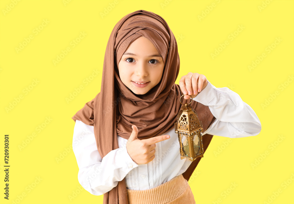 Little girl in hijab pointing at Muslim lantern on yellow background