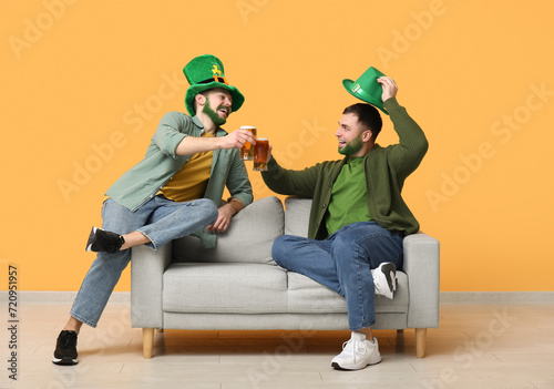 Young men in leprechaun hats with green beards holding glasses of beer and sitting on sofa near yellow wall. St. Patrick's Day celebration