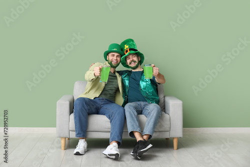 Young men in leprechaun hats with green beards holding glasses of beer and sitting on sofa near green wall. St. Patrick's Day celebration photo