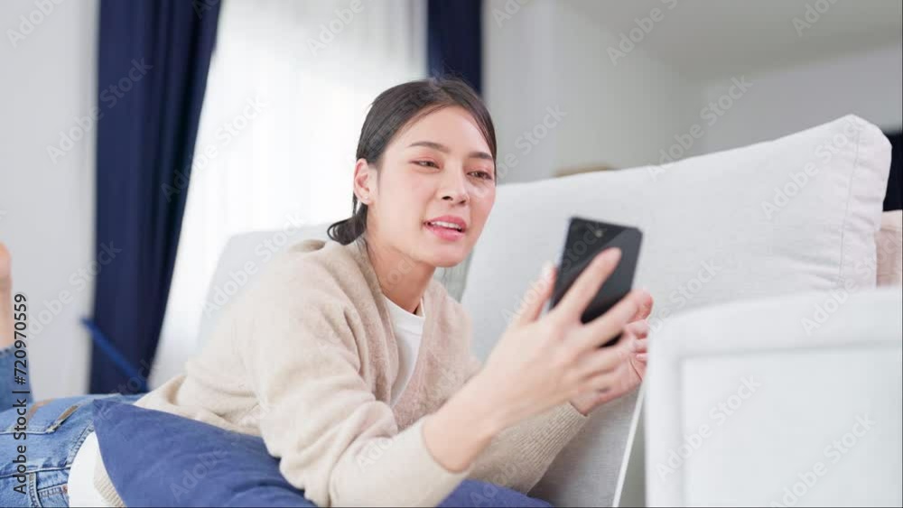 Happy young asian woman relaxing at home. Female smile lying on sofa and holding mobile smartphone. Girl using video call to friend Happy young asian woman relaxing at home. Female smile lying on sofa and holding mobile smartphone. Girl using video call to friend