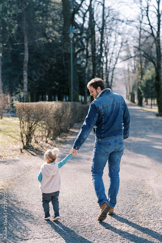 Dad with a little girl walk holding hands along the road in the spring park. Back view © Nadtochiy