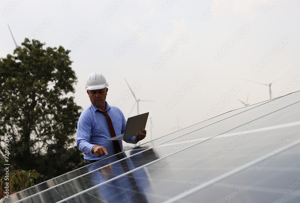 A senior male engineer inspects solar panels on the windmill farm. Clean energy. Engineers inspect a solar panel system with a turbine farm in the background, a renewable energy concept