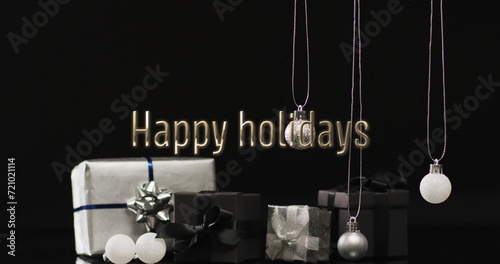 Happy holidays text over christmas presents and baubles hanging on dark background